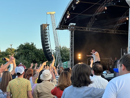 A stage is on the right hand side of the image, with a long speaker in the middle. A singer is stood on the stage, reaching out to the crowd. On the left hand side, the crowd has their hands in the air.