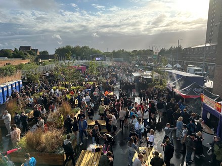 The camera is looking down to a crowd of people below. At the front of the shot are wooden picnic benches, with food stalls to the right hand side. Above the shot is a blue sky with lots of cloud.