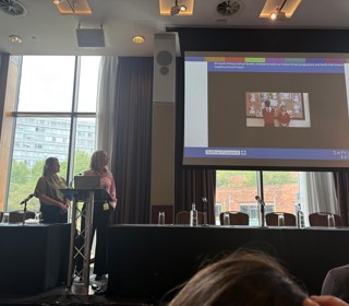 Two people on a stage, looking up at a presentation showing two pupils talking. 