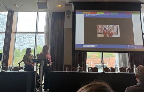 Two people on a stage, looking up at a presentation showing two pupils talking. 