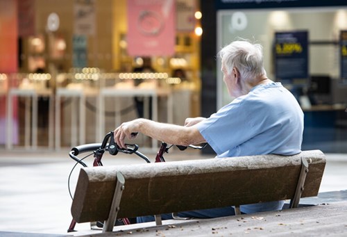 An older man sitting on a bench in town. He is wearing a smart blue t-shirt. His hands are resting on the handles of his wheel walker mobility aid