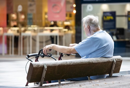 An older man sitting on a bench in town. He is wearing a smart blue t-shirt. His hands are resting on the handles of his wheel walker mobility aid