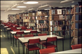 old photo of crosby library second floor with the left side having a line of tables and chairs and the right side having a line of metal bookshelves